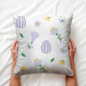 Poduszka trzymana dłońmi na jasnej pościeli, widoczny liliowy motyw kwiatowy