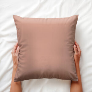 poduszka dekoracyjna w pudrowym różu trzymana dłońmi na białej pościeli, widok z góry