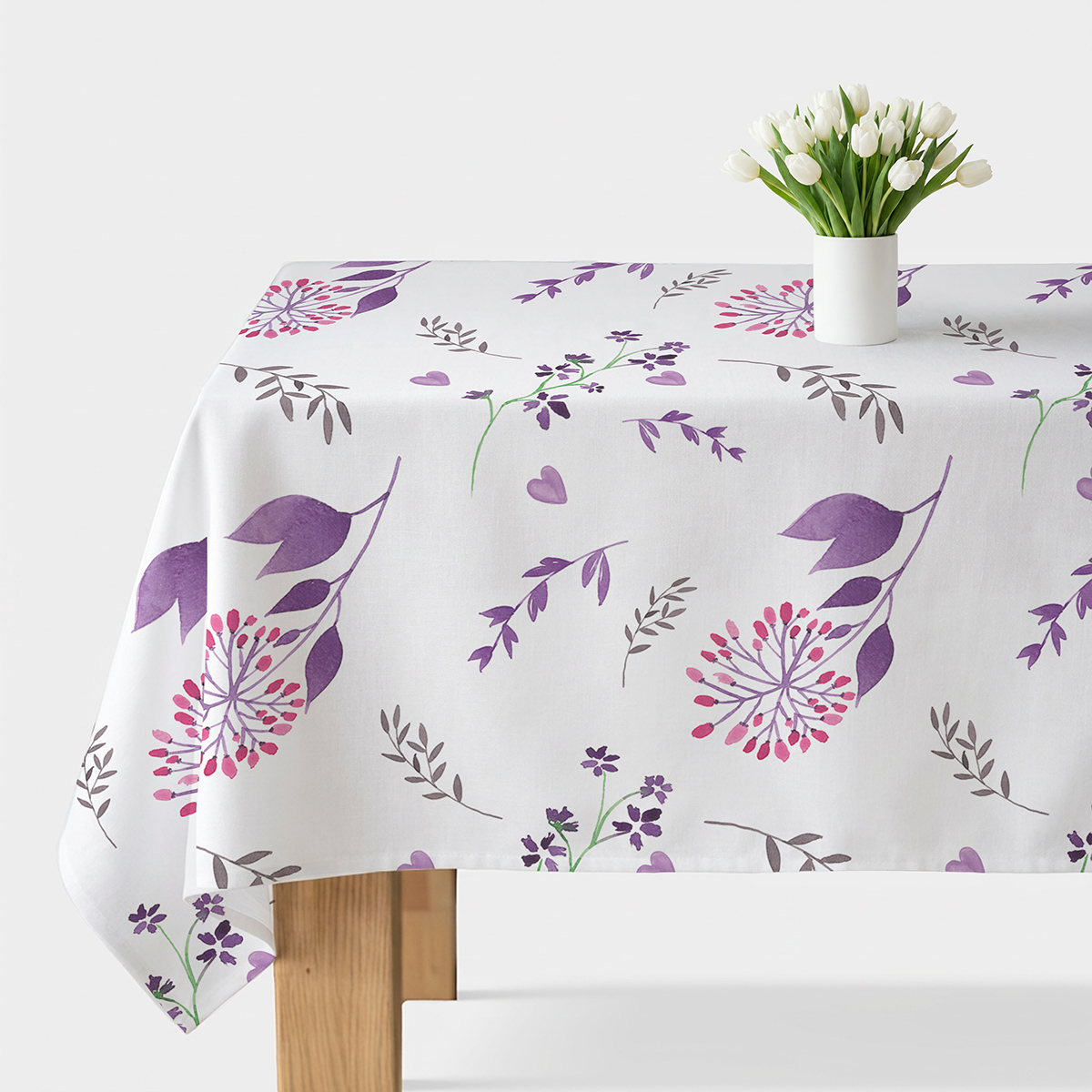 Stół nakryty obrusem z akwarelowym wzorem fioletowych gałązek i różowych kwiatów z białym wazonem z tulipanami