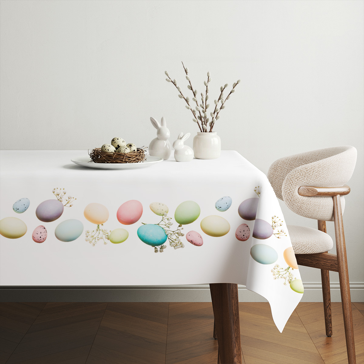 aranżacja stołu z obrusem ozdobionym pastelowymi jajkami, porcelanowymi zajączkami i gałązką bazi
