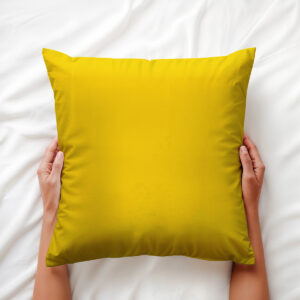 dłonie trzymające żółtą poduszkę na białej pościeli, ujęcie detalu