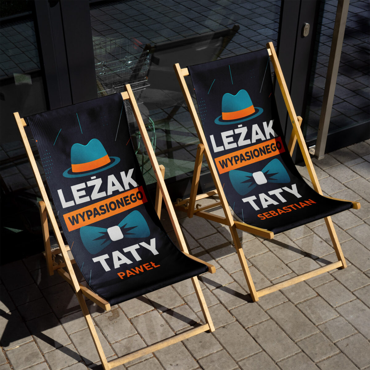 Zbliżenie na turkusowy kapelusz, pomarańczowy pasek i biały napis «LEŻAK» na granatowym materiale leżaka — close-up personalizacji imienia dla taty