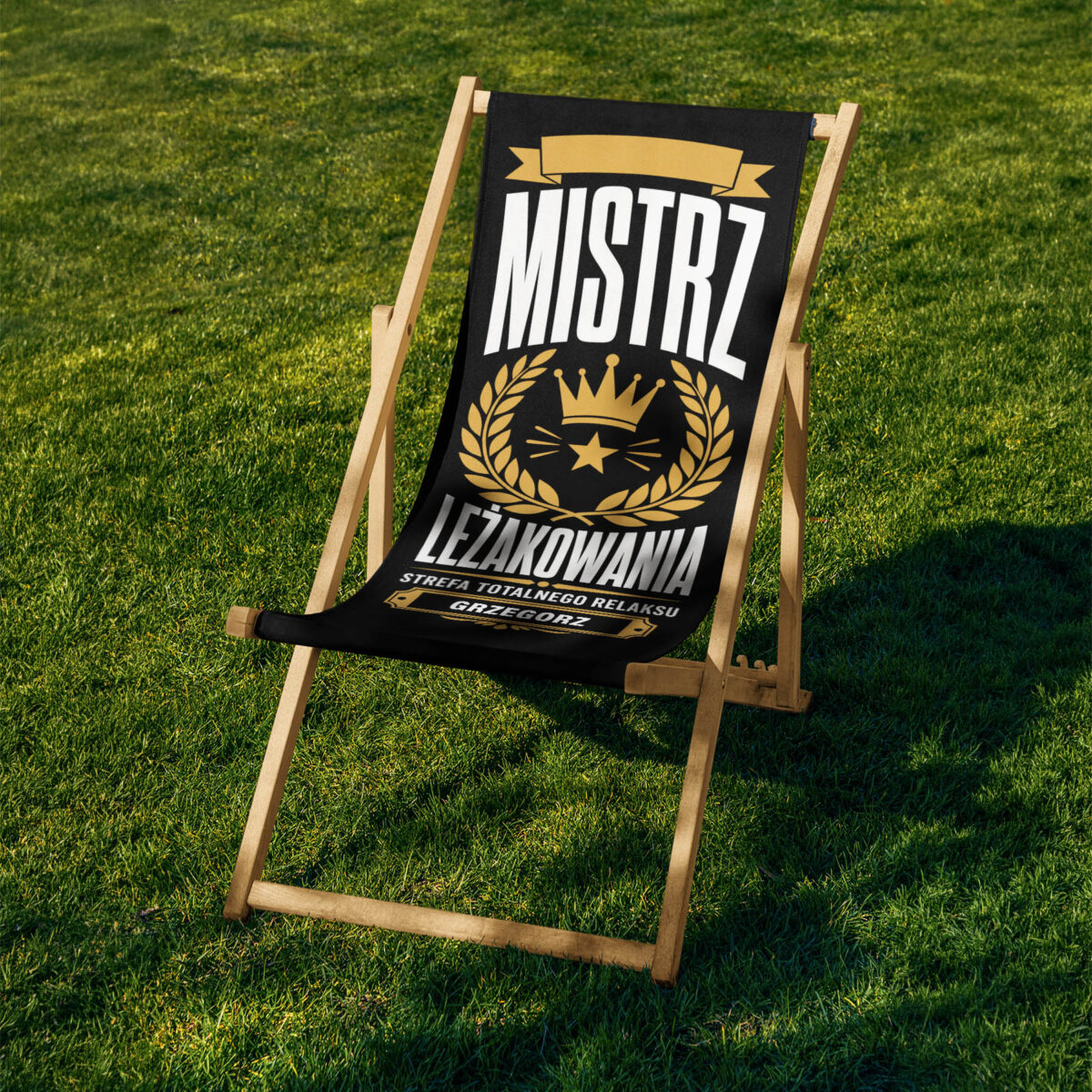 Pojedynczy leżak ustawiony na trawie z czarnym tłem nadruku i złotymi elementami oraz wyraźnym miejscem na personalizację imienia.